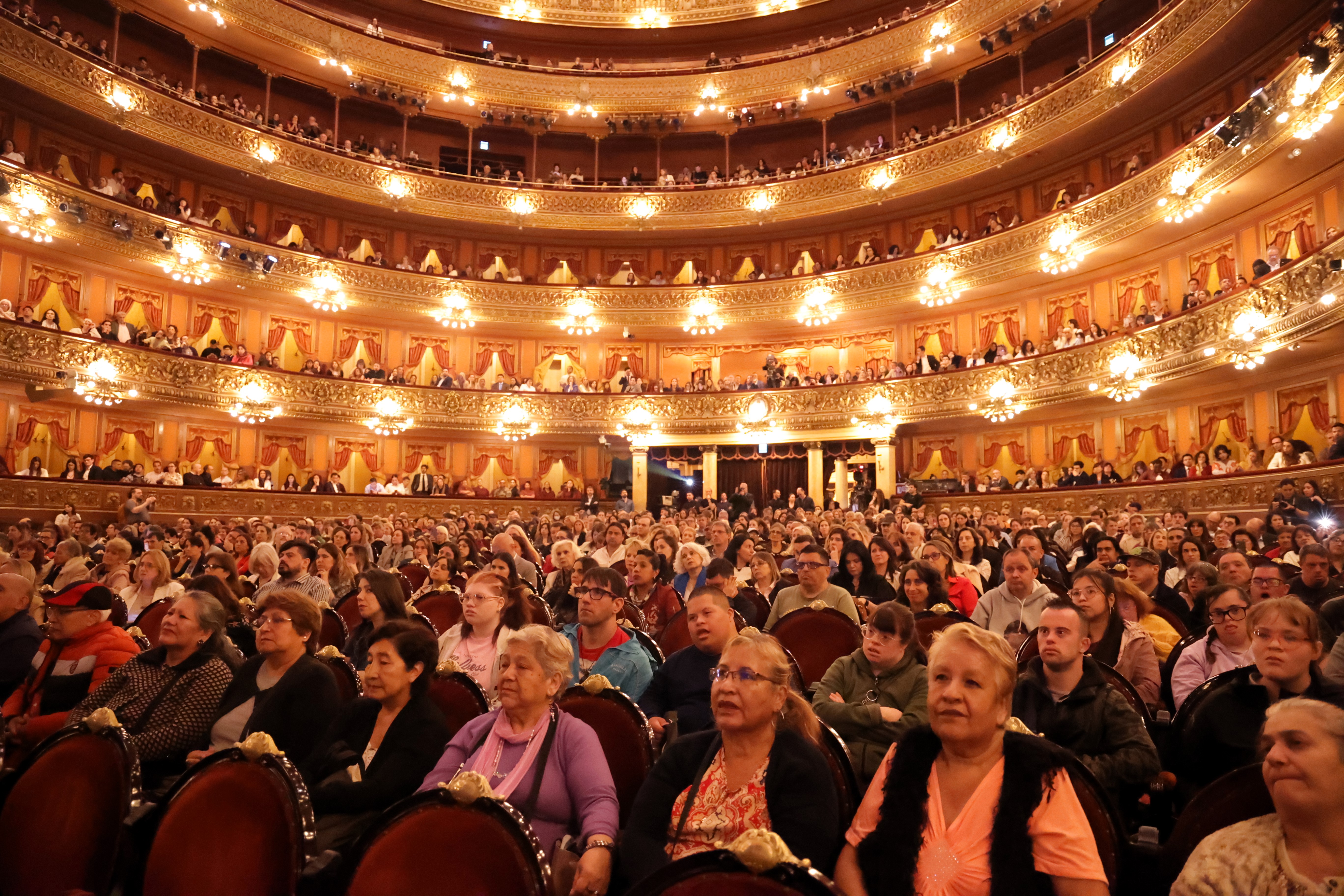 La imagen captura el vasto salón principal del Teatro Colón, lleno de espectadores. La sala es impresionante, con una arquitectura elaborada, decoraciones doradas y lámparas de luz cálida que iluminan el ambiente. Los asientos son de madera, con un diseño clásico, y parecen estar bien ocupados por una mezcla diversa de personas.  En el primer plano se pueden ver varias filas de público, con diferentes edades y expresiones. Algunas personas están atentas y observando lo que ocurre en el escenario, mientras que otras parecen conversar o reír. Entre ellos hay una notable diversidad, incluidos individuos con discapacidades visibles. En el fondo, las gradas se elevan en varias filas, llenas de asistentes que también están prestando atención al escenario.