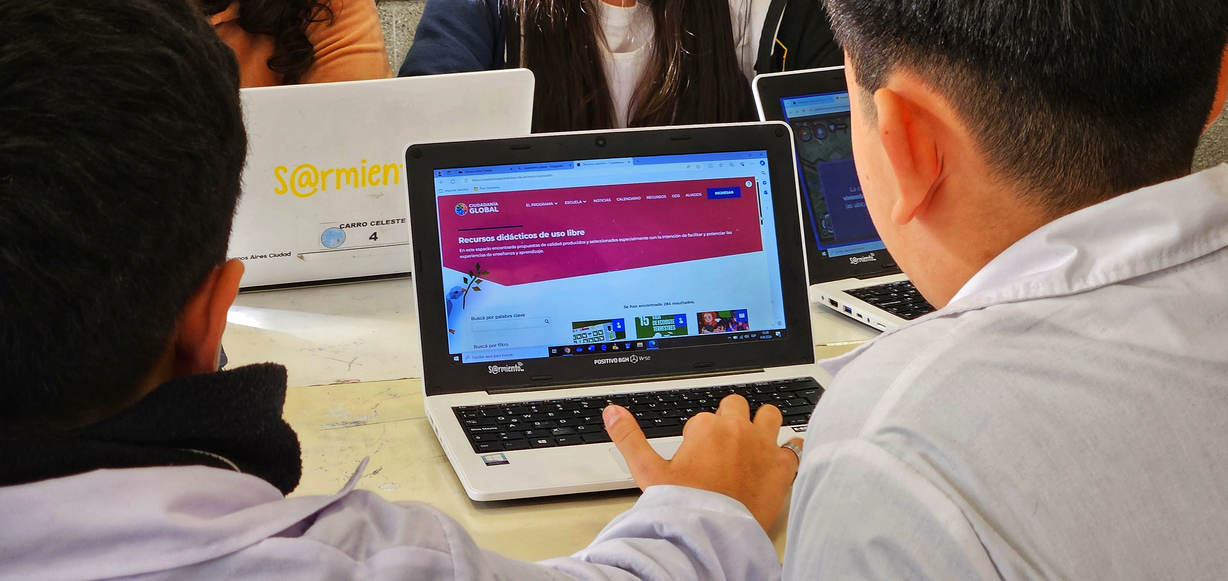 La imagen muestra a dos estudiantes de espaldas, sentados en un aula frente a sus computadoras portátiles. En la pantalla de la computadora más cercana, se puede ver una página web que ofrece recursos didácticos de uso libre, con un diseño colorido y accesible. Los estudiantes están enfocados en la pantalla, interactuando con el contenido en un ambiente de aprendizaje. Al fondo, hay otros compañeros, algunos de ellos visibles junto a las laptops. La escena sugiere un ambiente colaborativo y educativo, donde se fomenta el uso de la tecnología en el proceso de enseñanza.