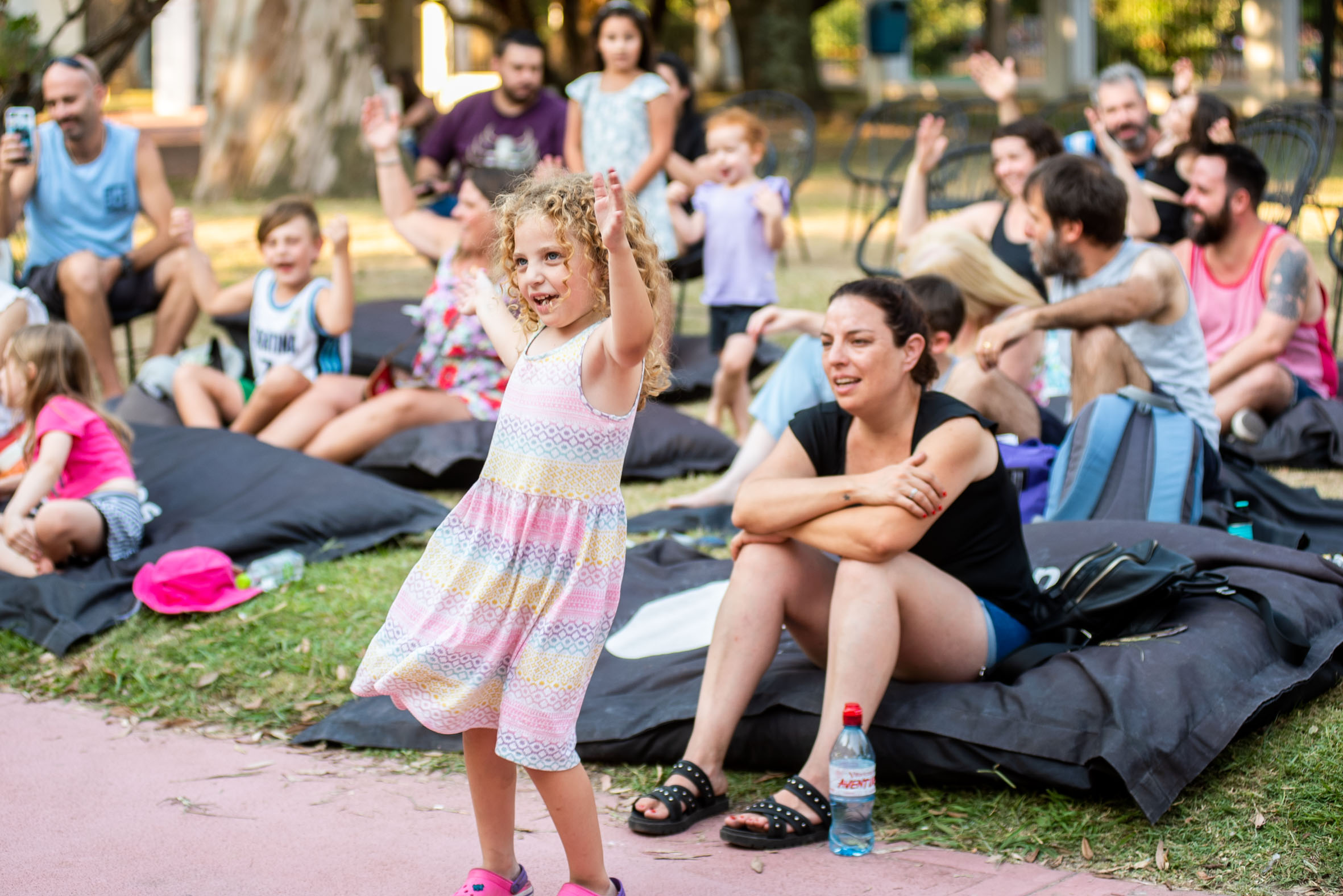 Imagen de una niña bailando al aire libre. De fondo se observan adultos sentados sobre colchonetas en el pasto