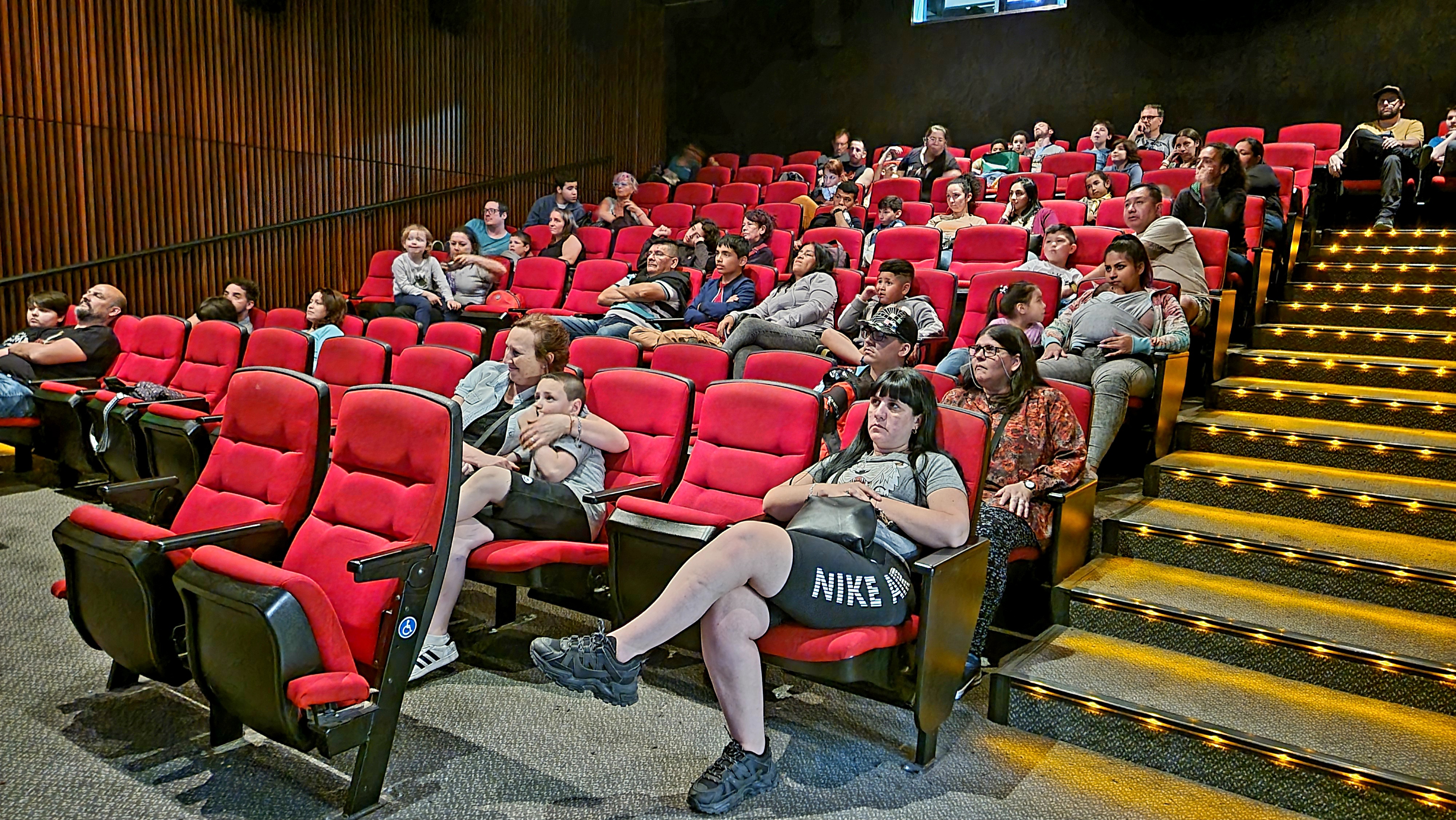 Fotografia tomada dentro de la sala donde se observan a los espectadores sentados y rescotados en las sillas observando el espectaculo