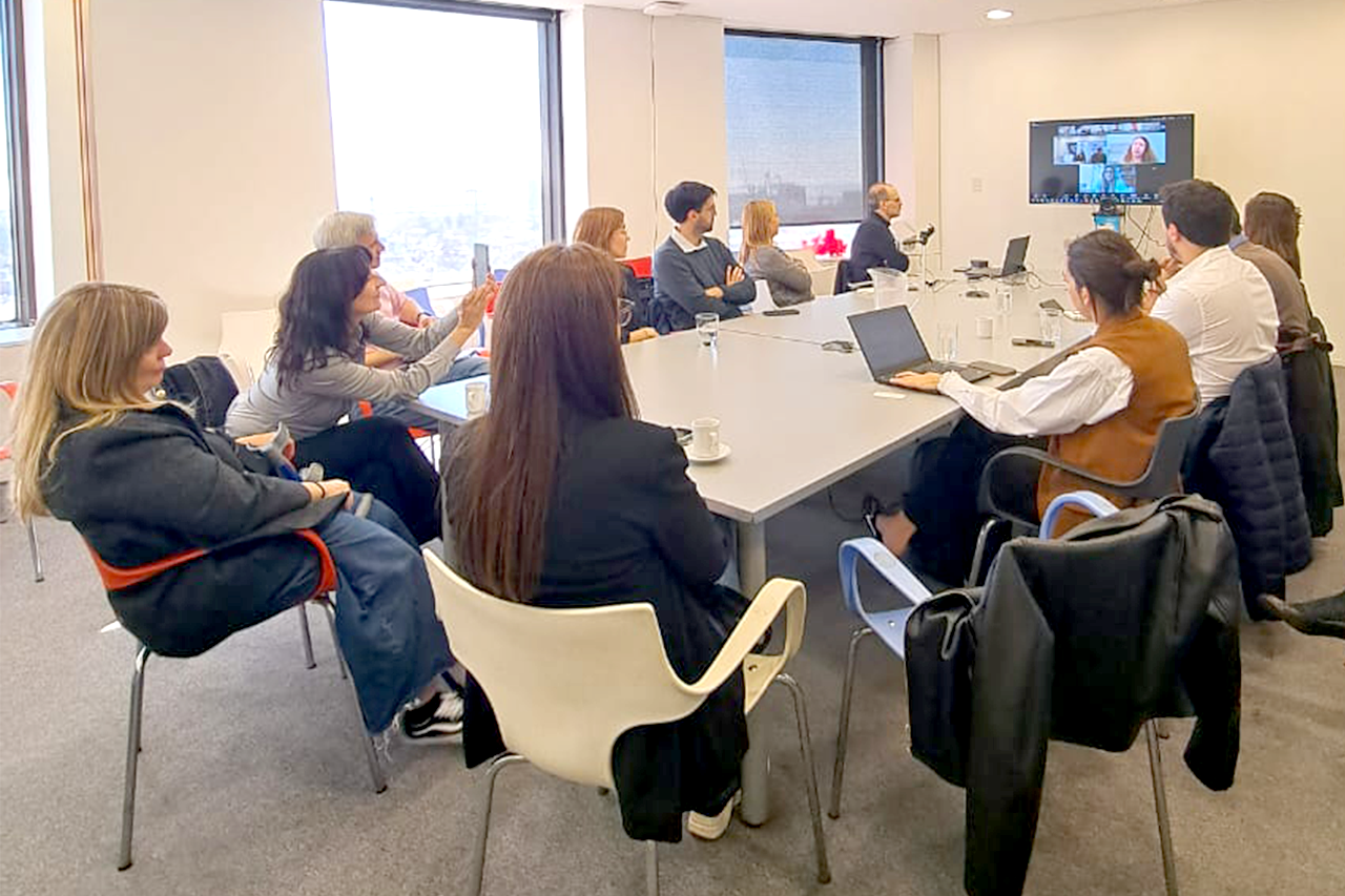 Un grupo de personas reunidas en una sala de oficina alrededor de una mesa, participando en una videollamada proyectada en una pantalla.