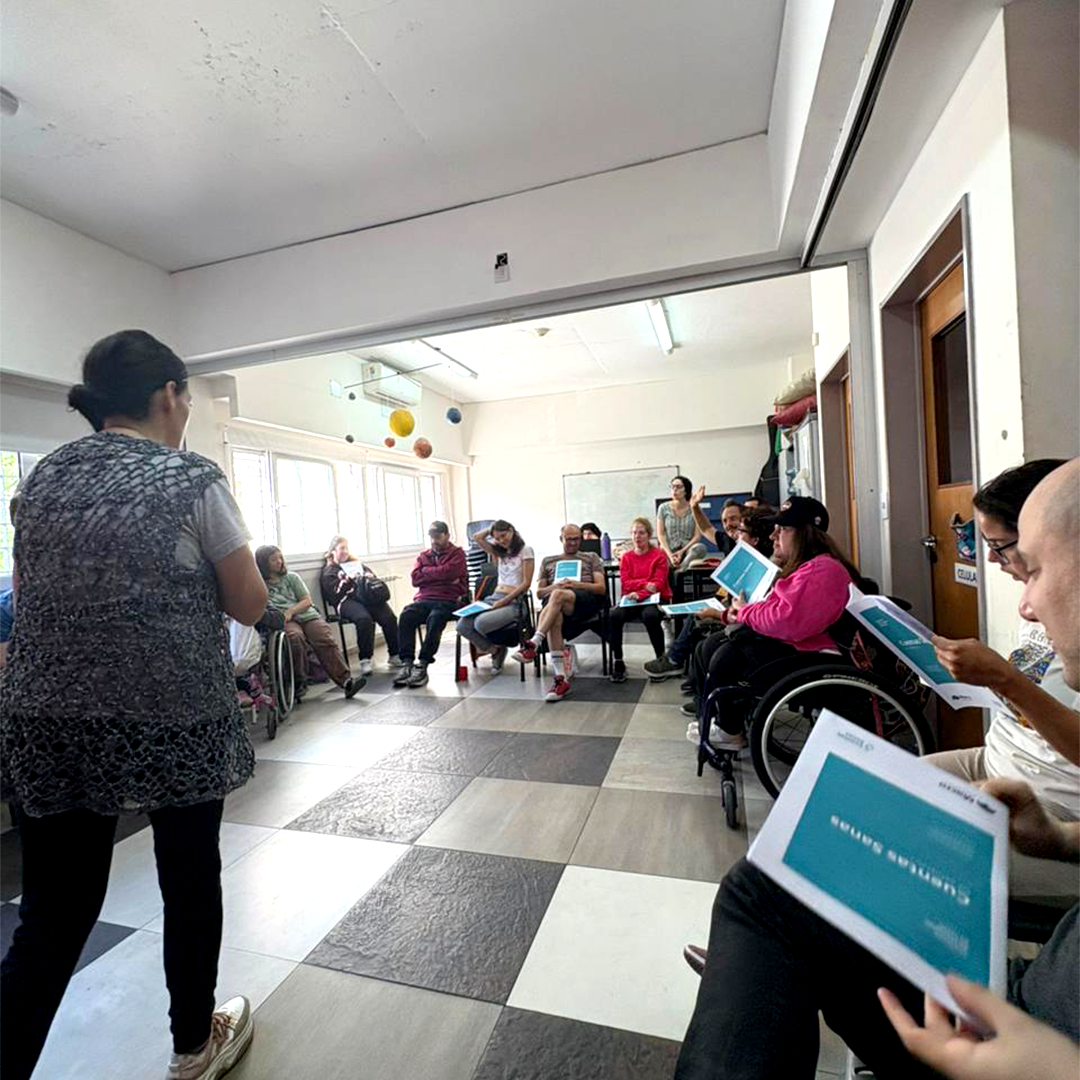 Personas sentadas en círculo en un salón con pisos a cuadros, participando del taller guiado por una facilitadora.