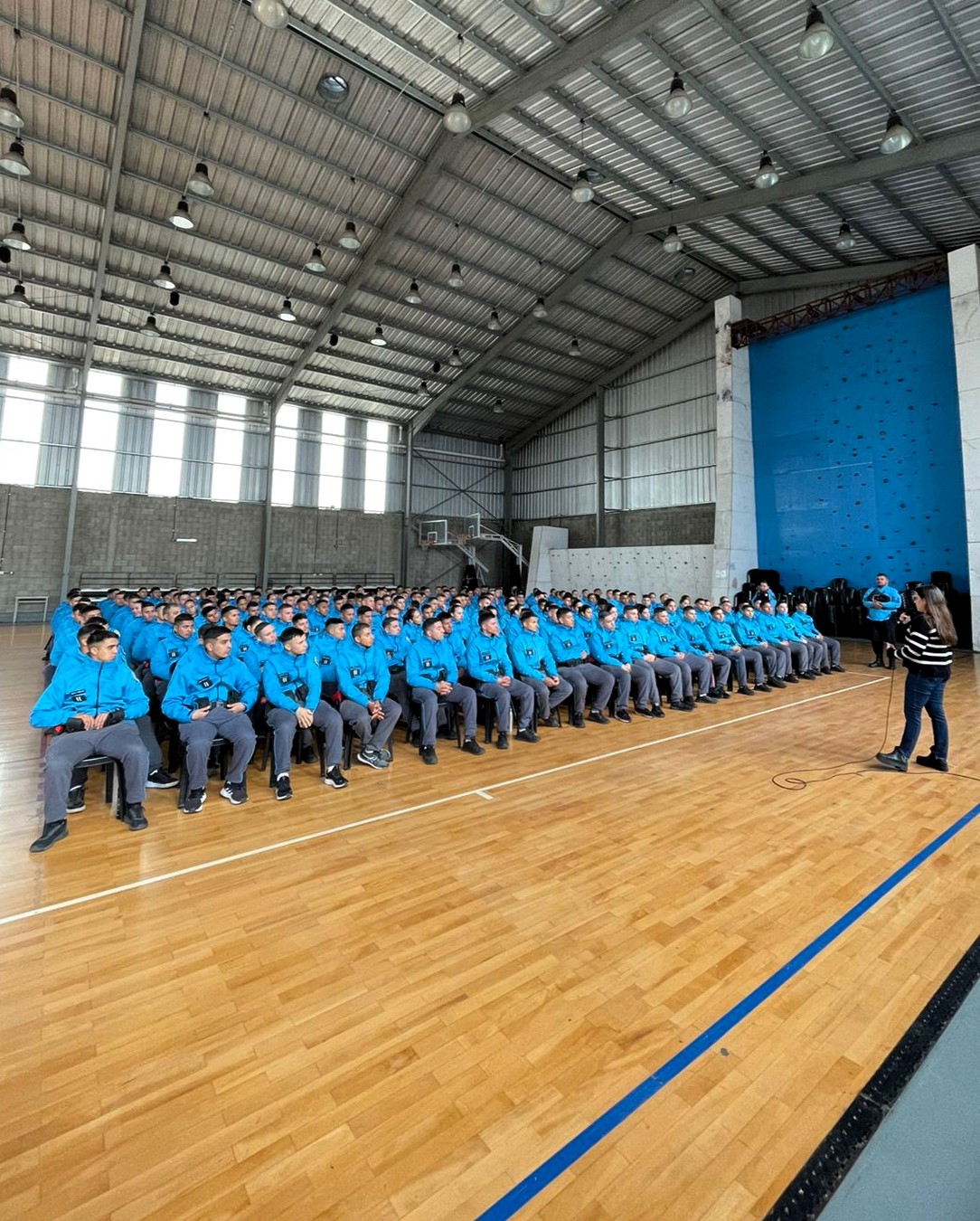 La imagen muestra un grupo de cadetes reunidos en un gimnasio o espacio deportivo. Están sentados en filas, todos vistiendo chaquetas azules, se persive un ambiente de formación. Al frente, hay una persona de pie, un instructor, quién se dirige a los cadetes. El espacio es amplio, con una pared de escalada visible al fondo, lo que indica que podría ser un lugar utilizado para diversas actividades físicas o de entrenamiento. La disposición ordenada de los cadetes sugiere un ambiente de atención y respeto hacia la actividad que se está llevando a cabo.