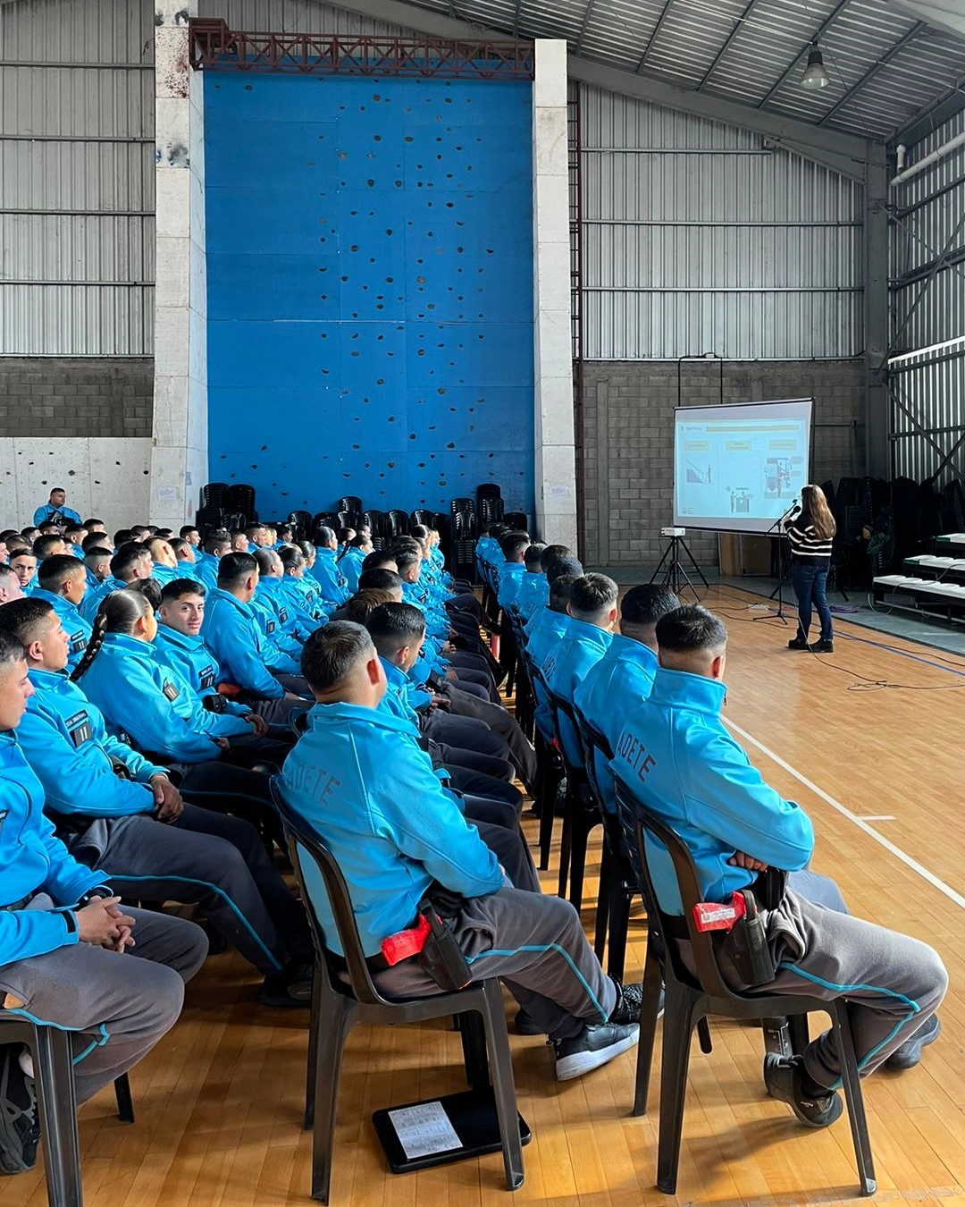 La imagen muestra un grupo de cadetes sentados en un gimnasio o espacio de entrenamiento. Están organizados en filas, todos vistiendo chaquetas azules, lo que indica que están en una sesión de capacitación. Al frente, hay una persona de pie, que está presentando información en una pantalla. La pared de escalada azul al fondo sugiere que el lugar se utiliza para diversas actividades físicas. La disposición ordenada de los cadetes refleja un ambiente de atención y respeto hacia la presentación que se está llevando a cabo. Además, algunos cadetes tienen un dispositivo o cuaderno en sus piernas, lo que indica que están tomando notas o siguiendo la presentación.