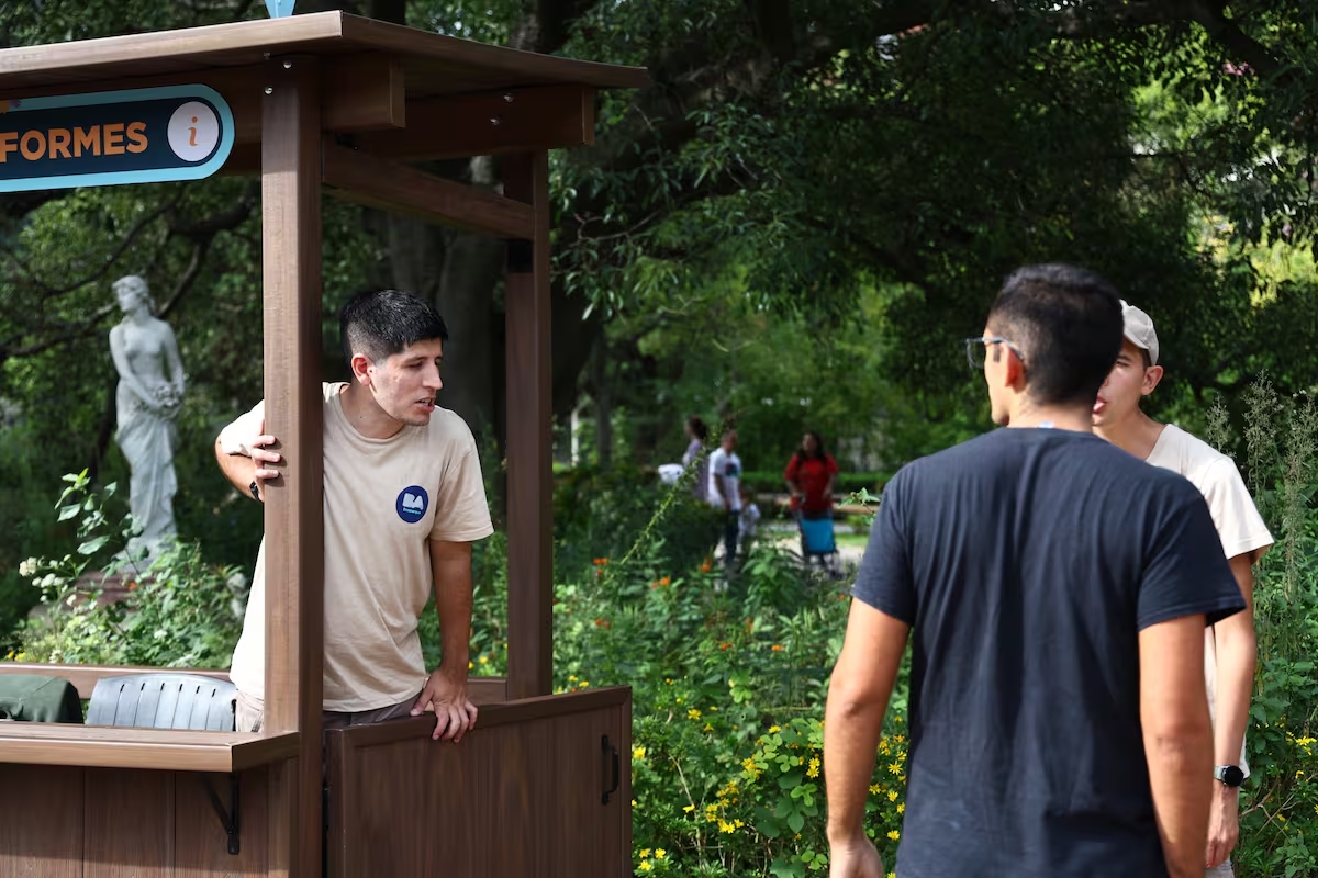Trabajador brinda información a visitantes en una posta del Ecoparque, en un entorno verde con senderos y público circulando.
