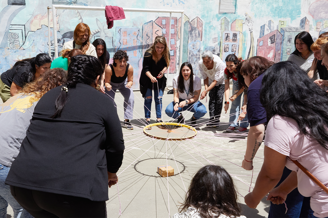 Actividad grupal durante el taller con personas en círculo sosteniendo cuerdas unidas a un aro central frente a un mural.