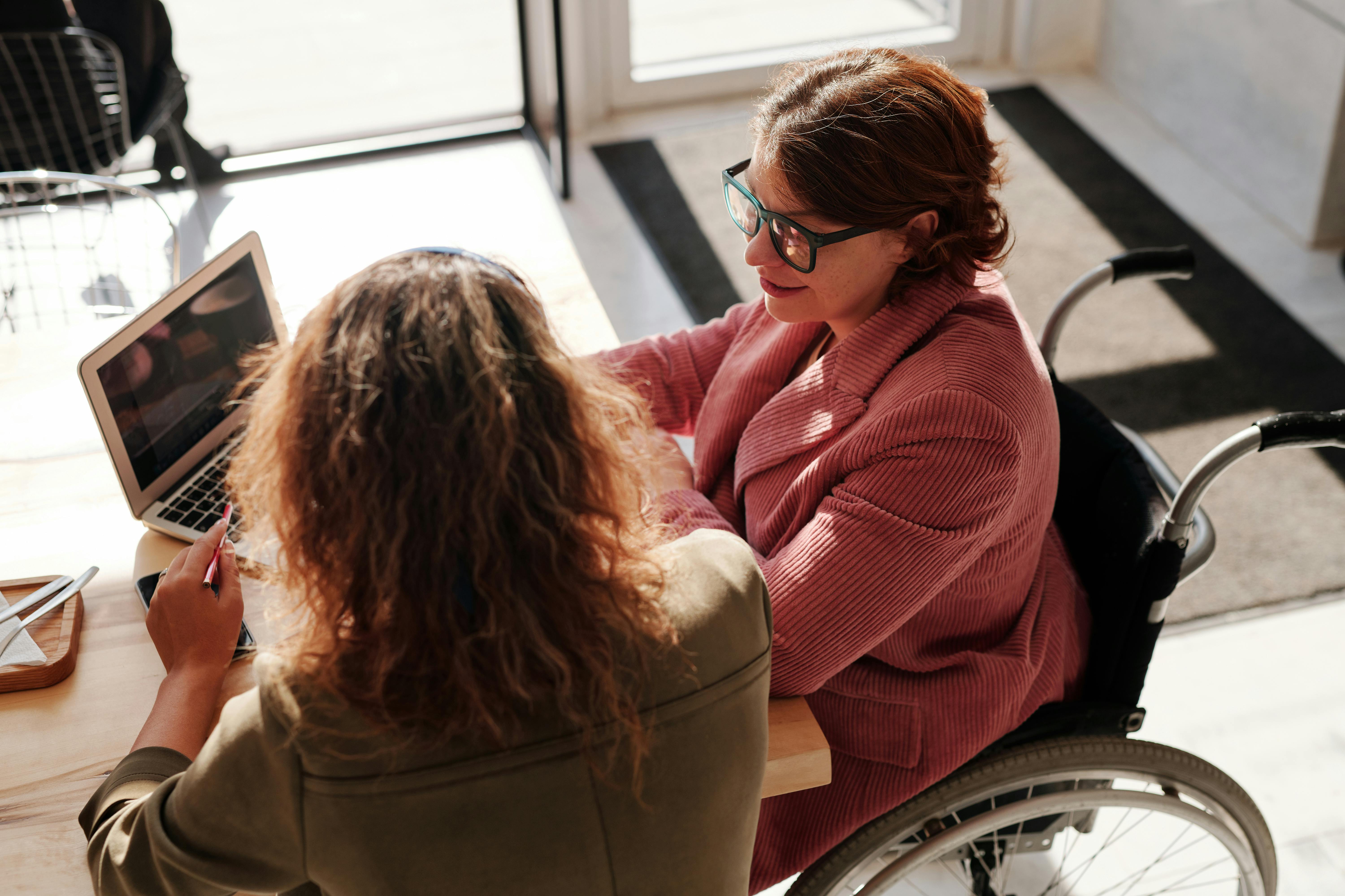 En la imagen se puede ver a dos mujeres en un entorno de oficina o trabajo colaborativo. Una de ellas está sentada en una silla de ruedas y lleva una chaqueta de color rosa claro. Ella está hablando y parece estar interesada en la conversación. La otra mujer, de cabello rizado y suelto, está sentada frente a ella y sostiene un bolígrafo, probablemente tomando notas en un cuaderno o en su dispositivo móvil, mientras observa atentamente a su interlocutora. Ambas parecen estar en una conversación animada, con un ambiente iluminado por la luz natural que entra por una ventana cercana. El mobiliario es moderno y acogedor, con una mesa de madera clara.