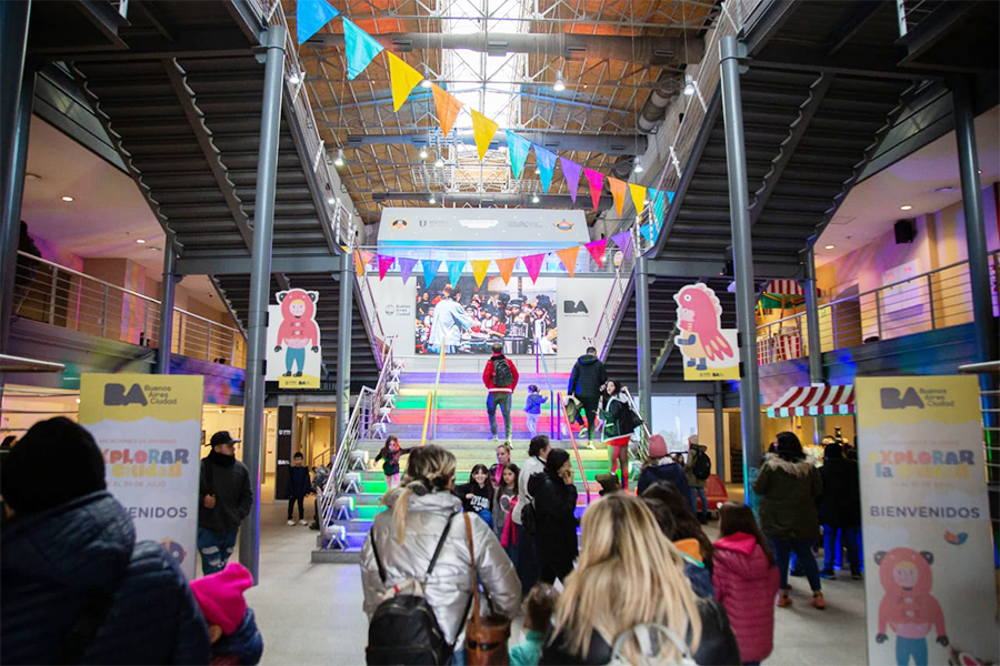 Ingreso a un centro cultural decorado con colores y banderines por las vacaciones de invierno. Personas recorren el espacio y suben una escalera iluminada.