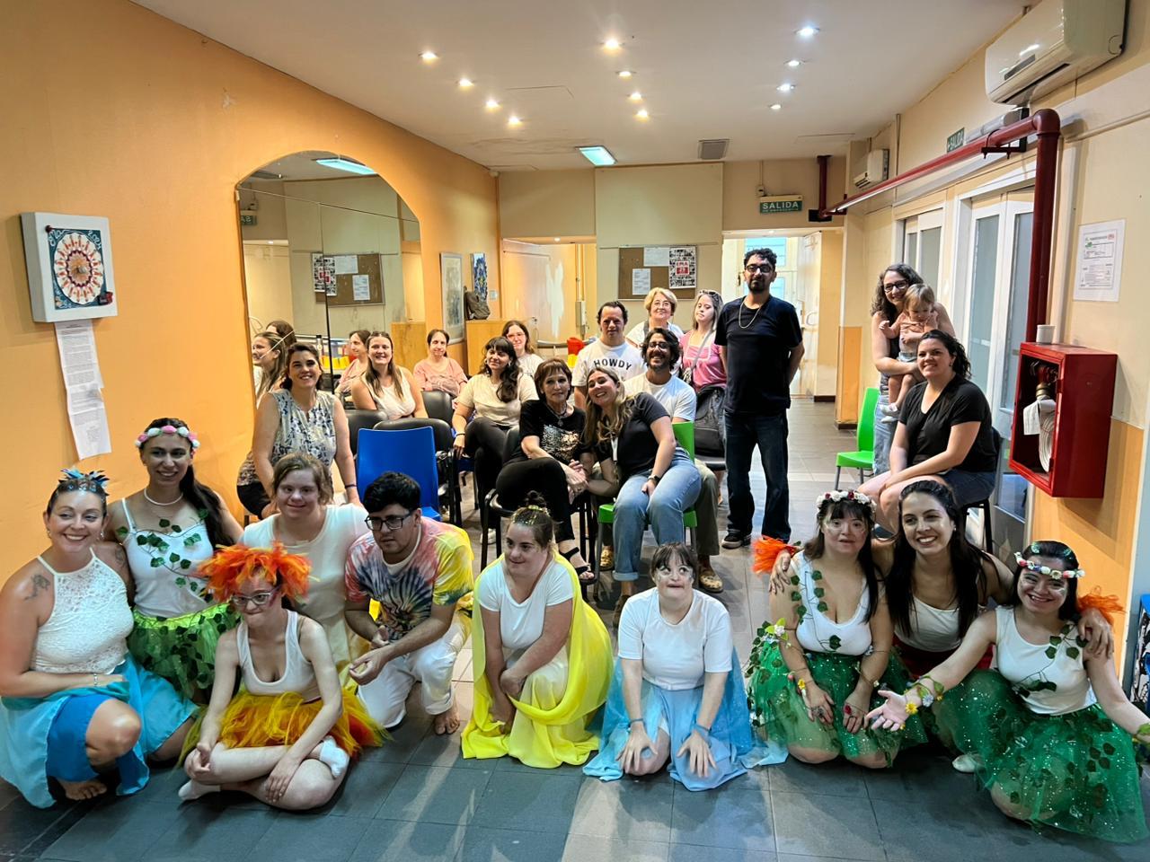 Grupo de artistas con vestuario de danza teatro durante una actividad junto a coordinadores y público de fondo.