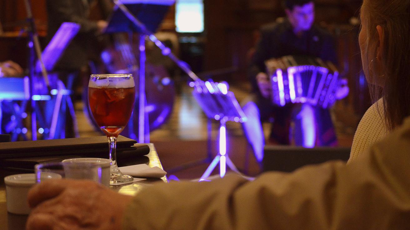 mano de una persona sobre una mesa. De fondo se ve un hombre con un bandoneon 