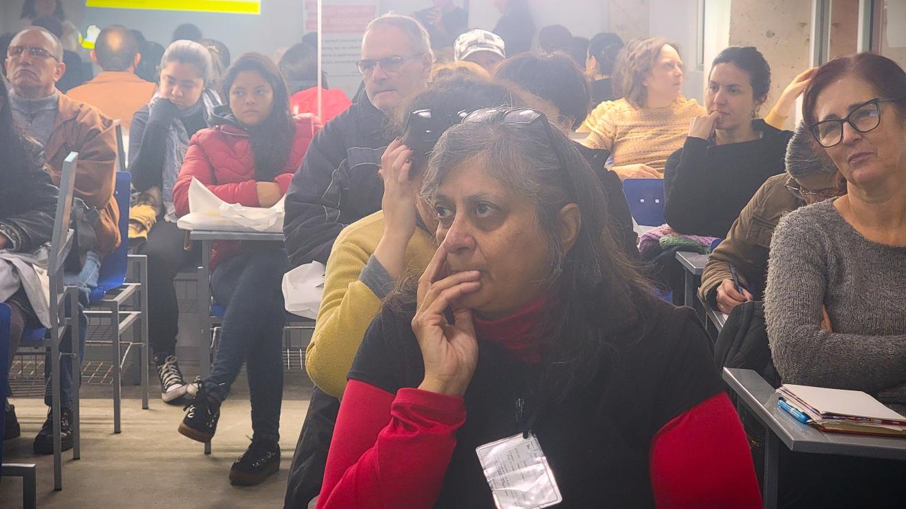 La imagen muestra a un grupo de personas sentadas en un aula. En el primer plano, se puede ver a una mujer con cabello largo y oscuro, que lleva una camiseta negra y un suéter rojo. Ella parece estar pensativa, con una mano en su boca. Al fondo, hay varias personas más, algunas de las cuales están mirando hacia adelante, mientras que otras parecen estar conversando. La atmósferae de atención y concentración