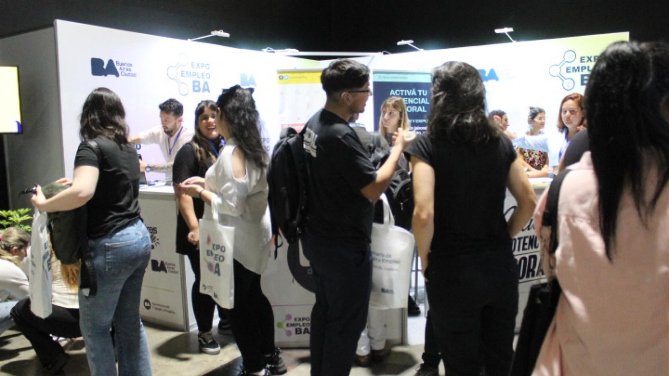 La imagen muestra un evento, una feria de empleo, titulado "Expo Empleo BA". En el fondo, se puede ver un stand con el logotipo de Buenos Aires y varios carteles que promueven la activación de potenciales laborales.  En primer plano, hay un grupo de personas, principalmente jóvenes, que parecen estar interactuando entre sí y con los representantes del stand. Algunos llevan bolsas de tela con el logo del evento. La atmósfera parece ser de networking y aprendizaje, con un enfoque en oportunidades