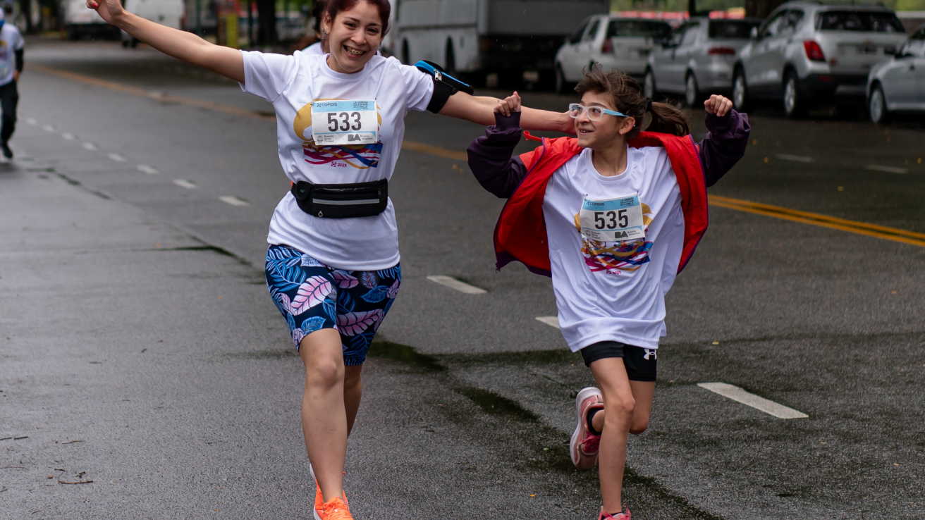 La imagen muestra a una mujer y una niña corriendo juntas en la carrera. Ambas llevan camisetas blancas con números de participante. La mujer, que tiene el cabello recogido, sonríe y parece estar disfrutando del momento, mientras que la niña, que lleva gafas, también sonríe y corre con entusiasmo. La mujer viste un pantalón corto con un diseño de hojas y zapatillas naranjas, mientras que la niña lleva una chaqueta roja sobre su camiseta. El ambiente es alegre y dinámico, con un camino mojado que su