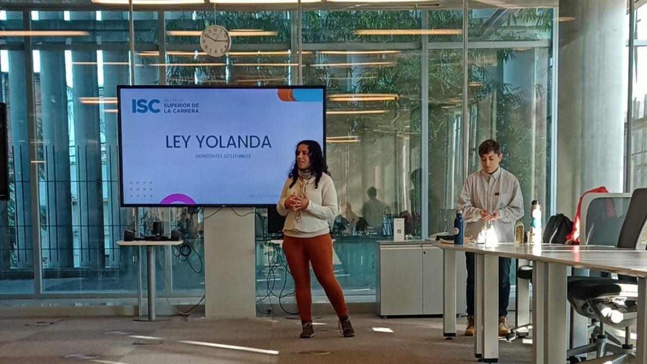 Dos personas de pie frente a una pantalla grande, hablandole a un publico que los esta escuchando.