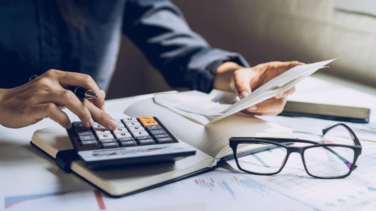 Se observan las manos de una persona, tomando con la mano izquierda un papel, y en su mano derecha utilizando una calculadora. Encima de la mesa hay papeles y unos lentes de ver.