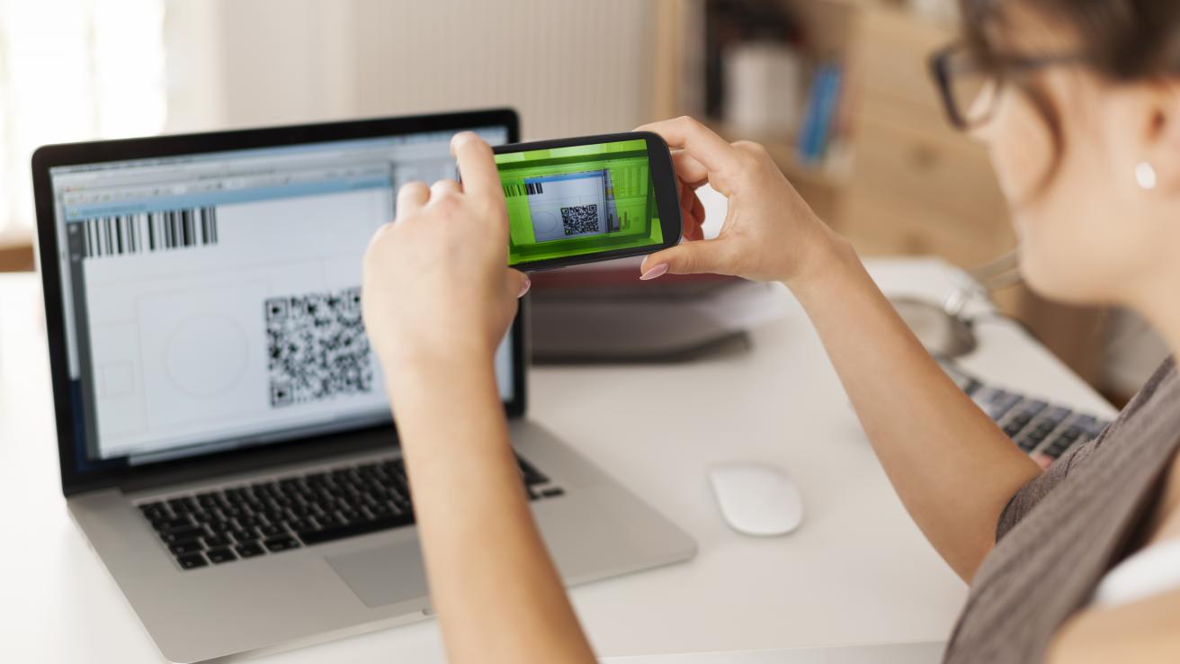 Una mujer escaneando el QR con su teléfono, el QR que muestra una pantalla de computadora 