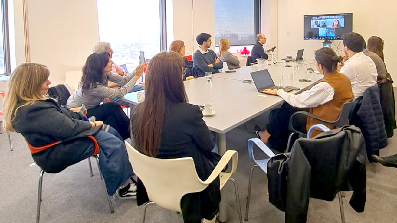 Un grupo de personas reunidas en una sala de oficina alrededor de una mesa, participando en una videollamada proyectada en una pantalla.