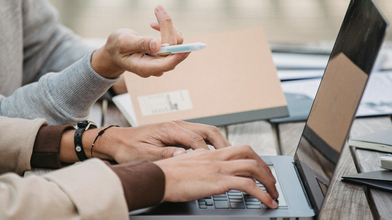 Dos personas colaborando en una computadora portátil al aire libre, con una de ellas escribiendo y la otra señalando con un bolígrafo sobre una mesa con cuadernos.