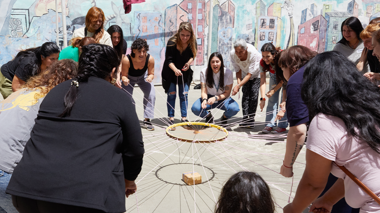 Actividad grupal durante el taller con personas en círculo sosteniendo cuerdas unidas a un aro central frente a un mural.