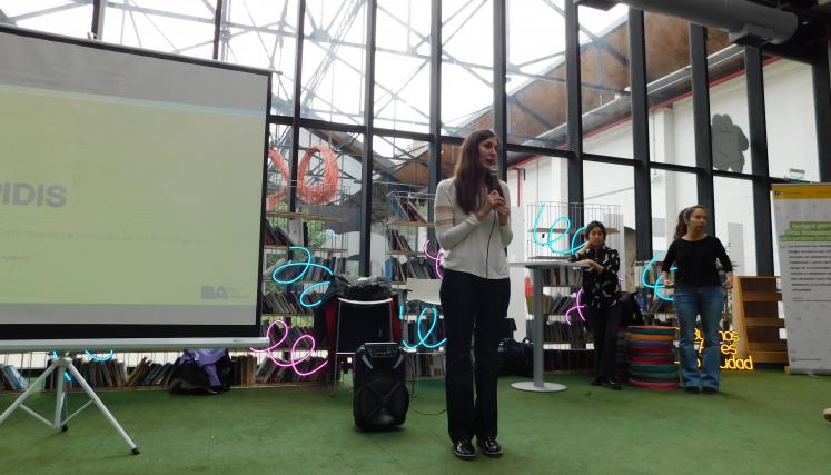 Fotografía en la que aparece Victoria Belotti, Gerente del Programa, dirigiéndose a los asistentes. A su lado derecho pantalla con la diapositiva de presentación. 