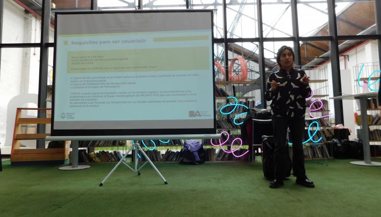 Fotografía en la que aparece una de las capacitadoras del programa dirigiéndose a los asistentes. A su lado derecho pantalla con la diapositiva de presentación. 