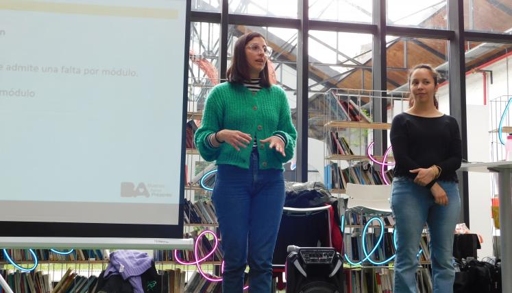 Fotografía en la que aparecen dos de las capacitadoras del programa dirigiéndose a los asistentes. A su lado derecho pantalla con la diapositiva de presentación. 