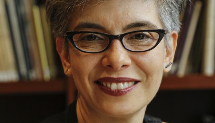 Retrato de María Laura Rosa, quien estará a cargo del conversatorio en el Museo de la Ciudad. Aparece de frente. Tiene el cabello corto y canoso, peinado con volumen, y usa anteojos oscuros de marco grueso. Sonríe levemente. Lleva puesta una musculosa negra y un collar plateado. De fondo, se ven estantes con libros.