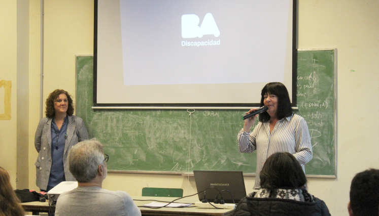 Expositora habla al público con micrófono, frente a una pantalla con el logo de BA Discapacidad, en un aula con pizarrón y material de apoyo.