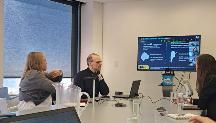 Personas reunidas en una sala de trabajo con laptops y materiales de apoyo, mientras siguen una videollamada proyectada en pantalla con una presentación sobre discapacidad y empleo.