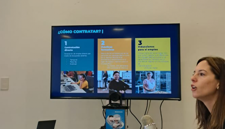 Mujer exponiendo en una reunión frente a una pantalla que muestra una presentación titulada '¿Cómo contratar?', con puntos sobre contratación directa, prácticas formativas e inducciones para el empleo