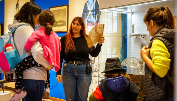 Imagen de una guía del museo con un abanico en la mano, conversando con una familia con dos niños