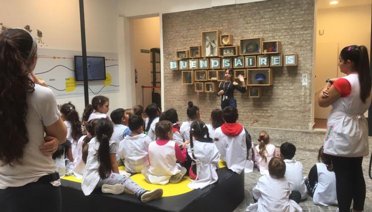 Recorrido guiado para escuelas en la exposición permanente del museo. En el patio central, un grupo de estudiantes sentados escucha con atención a la guía de sala, quien dicta el recorrido frente a una pared con objetos y la palabra “Buenos Aires”.