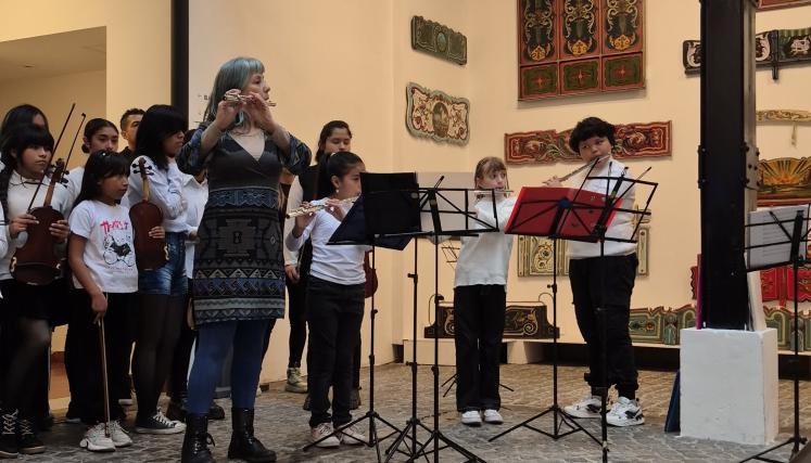 Una docente junto a niñas y niños interpretan una pieza con flautas traversas durante un concierto en el patio del Museo de la Ciudad, mientras otros integrantes de la orquesta esperan con sus instrumentos al fondo.